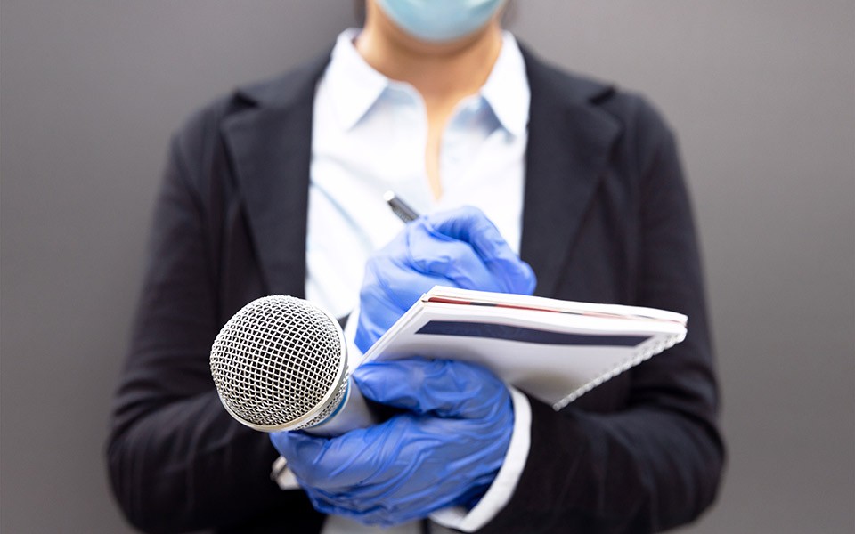 A woman with gloved hands and a face mask writes in a notebook and holds a microphone